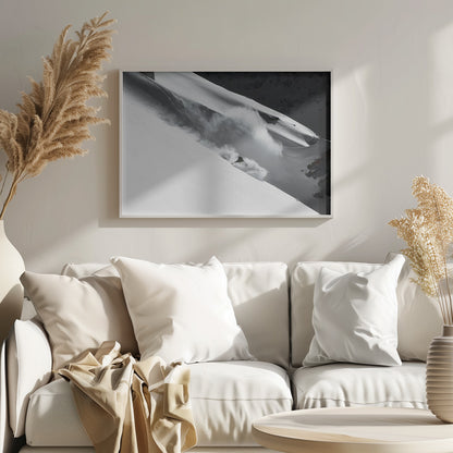 A dramatic black and white photograph of a skier speeding down a steep, snow-covered mountain, kicking up a large cloud of powder. The image is framed with a brushed silver border. Decor
