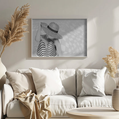 A black and white artistic photograph of a woman sitting down, her face obscured by a large, wide-brimmed sun hat with a black and white chevron pattern. She is wearing black and white striped leggings and holding the brim of her hat with one hand. The image is set against a plain background with patterned shadows and is presented within a silver frame. Artwork