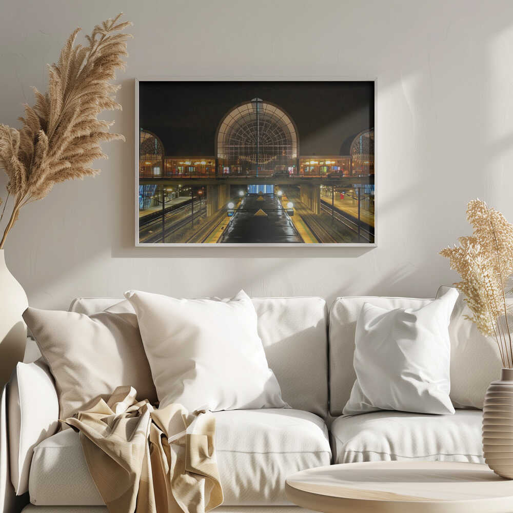 A symmetrical view of a modern train station at night, with a large illuminated arched glass window. Below, empty, wet train tracks and platforms reflect the station's lights, while cars cross an overpass in the middle ground. Wall Art