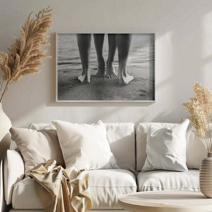 A framed, black and white photograph showing a low-angle view of two people's bare feet and lower legs standing on a wet sandy beach. The central pair of feet is in focus, while another person's legs in the foreground are blurred, creating depth. The water's edge is visible in the background. Artwork