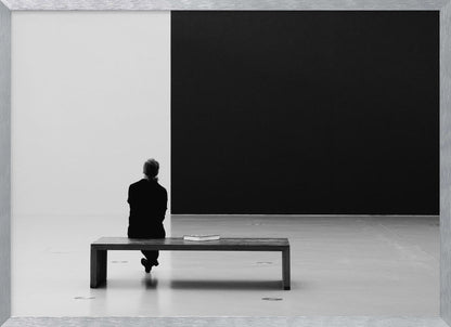 A black and white photograph of a person seen from behind, sitting alone on a wooden bench in a minimalist art gallery. They face a large, starkly divided wall, half white and half black. The scene is presented within a silver picture frame. Poster