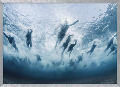 An underwater photograph looking up at a group of swimmers competing in a race. Their bodies are silhouetted against the bright surface of the turbulent blue water, captured mid-motion amidst a flurry of bubbles and foam. Print
