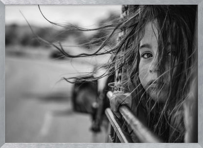 A close-up, black and white photograph of a young girl looking out of a moving vehicle. Her long hair is blowing wildly in the wind, partially obscuring her face as she peers intently at the camera with one eye. Her hand grips the edge of the open window. The background is blurred with motion, suggesting a journey. The photo is displayed in a silver frame. Poster