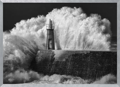 A dramatic black and white photograph of a massive wave crashing against a stone pier and a small lighthouse. The white foam of the wave towers over the structure, creating a powerful and dynamic scene against a dark sky, all enclosed in a silver frame. Poster