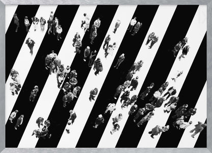 A striking black and white aerial photograph of dozens of people walking across a large, diagonally striped crosswalk. The high-contrast, graphic pattern of black and white stripes dominates the scene, with the small figures of pedestrians scattered across it, creating a sense of movement and urban anonymity. The image is presented within a simple silver frame. Print