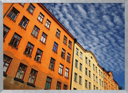 A low-angle shot of colorful buildings, predominantly a vibrant orange one, against a bright blue sky filled with a pattern of white, fluffy clouds. The image is presented within a silver-colored frame. Print