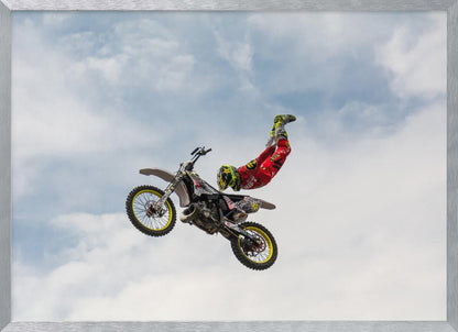 A motocross rider in red and yellow gear performs a high-flying stunt, arching their back while upside down in the air next to their dirt bike, set against a backdrop of a blue sky with white clouds and enclosed in a silver frame. Poster