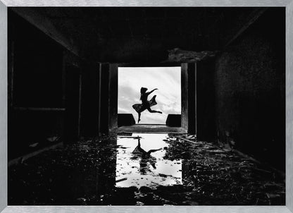 A dramatic black and white photo of a silhouetted dancer leaping in a bright doorway, viewed from inside a dark, derelict building. The dancer's reflection is captured in a puddle on the rubble-strewn floor. Poster