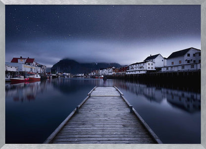 A long exposure photograph of a serene coastal town at night, viewed from a wooden pier that leads into calm, dark water. A lone person stands at the end of the pier, gazing at the town's reflected lights. Behind the village, a mountain looms under a breathtaking, star-filled sky. The image is presented within a silver-colored frame. Artwork