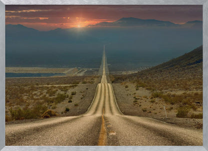 A high-angle view of a long, straight, and hilly road disappearing into the horizon of a vast desert valley at sunset, with mountains in the background under a warm, glowing sky, all enclosed in a silver frame. Poster