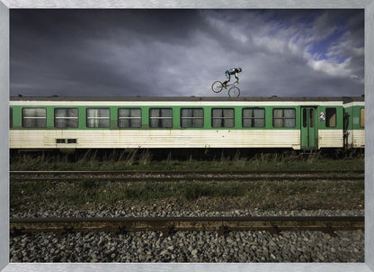 A BMX rider is captured in mid-air performing a stunt on the roof of a green and white passenger train car, set against a dramatic, stormy sky. The scene takes place on railway tracks with gravel and overgrown grass, and the entire image is enclosed in a silver frame. Decor
