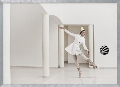 A ballerina dressed in a white quilted coat and a pointed hat dances en pointe in a minimalist white hall with large columns. She gracefully extends her arms, looking towards a black and white striped ball that appears to be floating in mid-air. The scene is presented within a silver picture frame. Wall Art