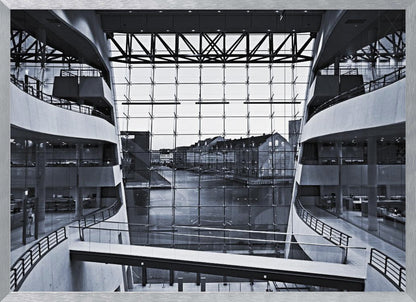 A symmetrical, black and white photograph taken from inside a modern building with curved balconies, looking out through a massive glass wall onto a canal and historic buildings on the opposite bank. A pedestrian bridge crosses the lower part of the frame. Decor