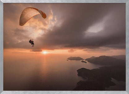 A scenic aerial view of a person paragliding over the ocean during a dramatic sunset. The orange and white parachute is in the upper left corner against a cloudy sky. The sun on the horizon casts a warm glow on the water, with silhouetted islands in the distance. Poster