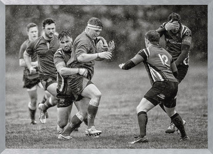 A dynamic black and white photograph of a rugby match being played in the rain. One player charges forward with the ball while an opponent wraps his arms around him in a tackle. Other players are in motion around them, capturing the intensity and physicality of the sport. Decor