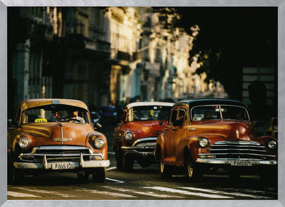 Three vintage American cars drive down a sunlit city street. The foreground features an orange taxi, with a red car and another orange vehicle behind it, all under the warm glow of late afternoon light casting long shadows. Print