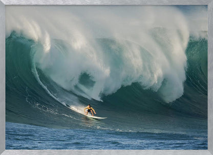 A lone surfer in a yellow shirt rides down the face of an enormous, powerful ocean wave as it crests into white foam, viewed within a silver picture frame. Wall Art