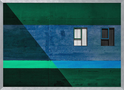 A framed abstract architectural photograph featuring a color-blocked facade. A central horizontal band of textured blue wall contains two small, distinct windows. This is flanked by upper and lower sections of dark green. A large, dark diagonal shape overlays the left side of the composition, creating sharp geometric lines. Decor