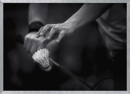 A dramatic black and white close-up photograph of a badminton player preparing to serve. The focus is on the player's hands, with one hand delicately positioned above a shuttlecock which is balanced on the strings of a racquet. Artwork