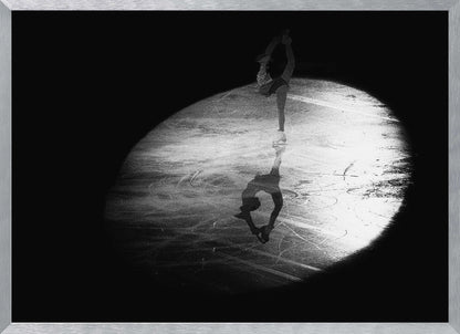 A dramatic black and white framed photograph of a female figure skater performing on ice. She is illuminated by a single spotlight, holding a graceful standing split pose, with her reflection visible on the ice below. Poster