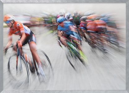 An action photograph of a professional cycling race captured with a prominent motion blur effect that conveys a strong sense of speed. A cyclist in an orange and black uniform is in the foreground, leaning into a turn, while a group of other riders in blue and multi-colored jerseys follows closely behind, their forms streaked and blurred. The entire scene is encased in a brushed silver frame. Poster