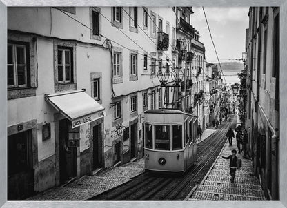 A black and white photograph of a vintage tram on a steep, narrow cobblestone street in an old European city. The street is lined with historic buildings and people are walking alongside the tracks. In the distance, a body of water is visible. Poster