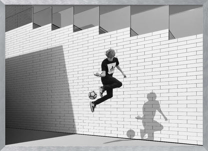 A framed, black and white photograph of a young person in mid-air, skillfully juggling a soccer ball with their foot against a clean, white brick wall. The person's shadow is cast sharply on the wall, mirroring the action, under a modern building with angled windows. Wall Art