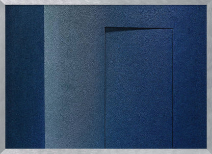 An abstract geometric artwork featuring textured panels in various shades of deep and dusty blue. The composition is divided into vertical sections, with a central rectangular shape that appears slightly recessed, creating a strong shadow line at its top edge. The piece is enclosed in a simple, brushed silver frame. Wall Art
