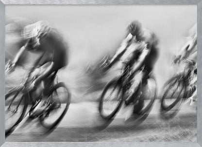 A dynamic black and white action photograph of three cyclists racing, captured with a slow shutter speed to create a dramatic motion blur effect. The riders and their bikes are blurred into streaks of light and shadow, conveying a powerful sense of speed and intensity. The image is presented within a silver-toned frame. Poster
