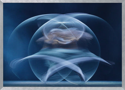 An abstract long-exposure photograph of a spinning dancer, creating ethereal, intersecting trails of blue and white light that form a glowing, orb-like shape against a deep blue background. The image is presented within a silver frame. Poster