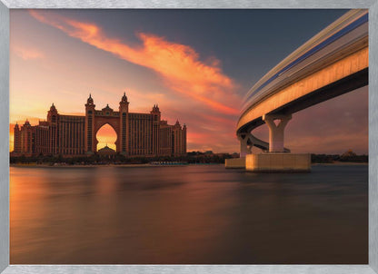 A scenic view of the Atlantis The Palm hotel in Dubai at sunset, with its grand architecture silhouetted against a vibrant orange and pink sky. A modern monorail track curves over the water on the right, with a blurred train indicating motion. The calm water in the foreground reflects the warm colors of the dusk sky. Decor