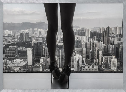 A framed black and white photograph showing the back of a woman's legs and high heels as she stands before a large window, looking out over a vast, dense cityscape with distant mountains under a cloudy sky. Artwork