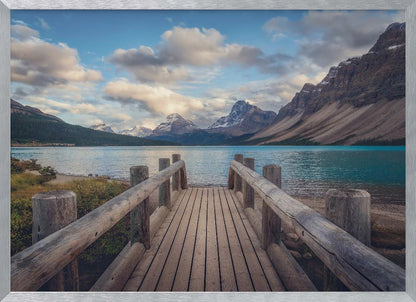 A wooden pier leads out onto a vibrant turquoise glacial lake, surrounded by towering, snow-dusted mountains under a dramatic cloudy sky. The image is presented in a silver frame. Artwork