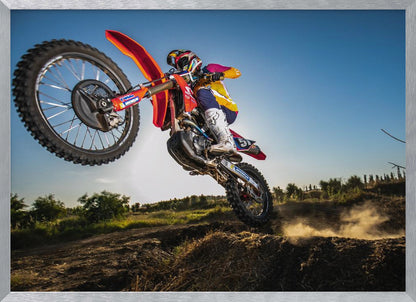 A dynamic, low-angle shot of a motocross rider in mid-air, jumping over a dirt mound. The rider is on a red, white, and blue dirt bike, wearing colorful racing gear and a helmet. The bike's front wheel is high in the air against a clear blue sky, while the back wheel is just clearing the jump, kicking up a cloud of dust. The background features a green, rural landscape. Wall Art