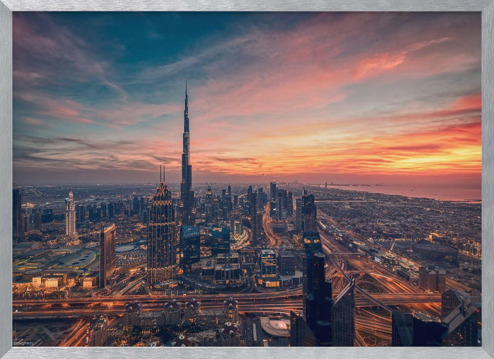 An aerial view of the Dubai skyline at sunset, with the Burj Khalifa prominently featured against a dramatic sky filled with orange, pink, and blue clouds. The sprawling city below is illuminated with golden lights from buildings and busy highways. Decor