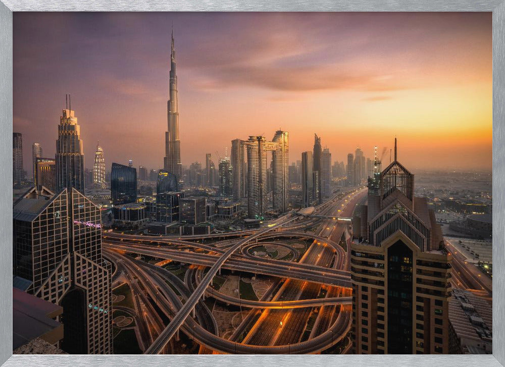 An elevated photograph of the Dubai skyline at sunset, featuring the towering Burj Khalifa against a hazy orange and purple sky. In the foreground, a complex network of illuminated highways and overpasses weaves between modern skyscrapers. Wall Art