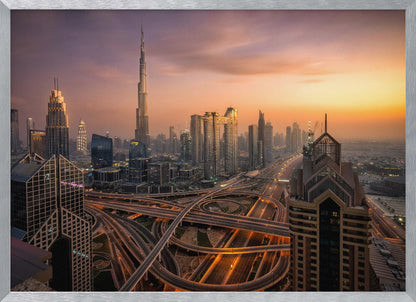 An elevated photograph of the Dubai skyline at sunset, featuring the towering Burj Khalifa against a hazy orange and purple sky. In the foreground, a complex network of illuminated highways and overpasses weaves between modern skyscrapers. Wall Art
