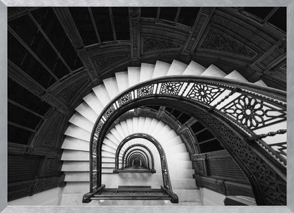A mesmerizing black and white photograph shot from a high angle looking down into the center of a spiral staircase. The bright white marble steps contrast with the dark, ornate metal railing, creating a repeating arched pattern that draws the eye downward into a seemingly endless tunnel. The image is presented within a silver-colored frame. Artwork