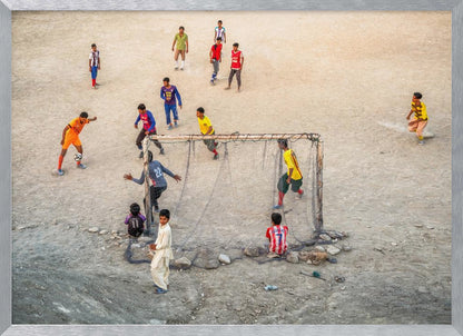 An overhead view of a group of boys and young men playing a spirited game of soccer on a large, dusty dirt field. A player in a bright orange uniform prepares to kick the ball towards a makeshift goal, defended by a goalie in a dark blue jersey. Other players in various colored shirts are scattered across the field, while a few young spectators watch from the sidelines. The entire image is enclosed in a silver frame. Wall Art
