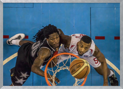 An overhead, bird's-eye view photograph of two basketball players looking up from below the hoop as a basketball goes through the net. The court is bright blue, and the image is enclosed in a silver frame. Poster