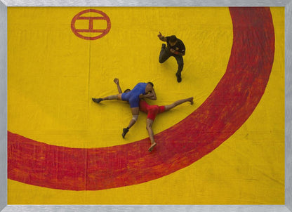 An aerial, high-angle photograph of a wrestling match in a silver frame. Two wrestlers, one in a blue singlet and the other in red, are locked in a hold on a vibrant yellow mat marked with a thick, painted red circle. A referee dressed in black stands nearby, gesturing. Artwork