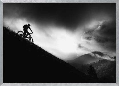 A striking black and white photo of a mountain biker's silhouette on a steep decline, set against a dramatic, light-filled cloudy sky with distant, misty mountains. Artwork