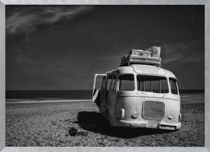 A dramatic black and white photograph of a vintage bus with luggage stacked on the roof, parked on a sandy beach. The ocean stretches out to the horizon under a dark, moody sky, with a small flock of birds flying in the distance. Print