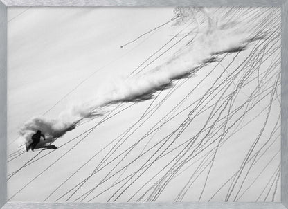 A dramatic black and white overhead photograph of a skier carving down a steep, powder-filled mountain slope, kicking up a huge plume of snow. Numerous ski tracks crisscross the pristine white snow, creating a pattern of dark lines. Poster
