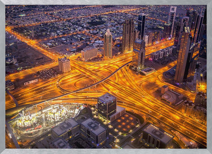 A stunning aerial view of a futuristic city at night, with brilliantly lit highways forming intricate patterns of golden light amidst towering skyscrapers, enclosed in a silver frame. Poster
