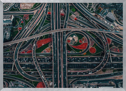 An aerial, top-down photograph of a sprawling and complex highway interchange, framed in silver. Multiple levels of roads and overpasses curve and intertwine, with blurred streaks of traffic visible. The spaces between the highways feature manicured green lawns and artistic red and patterned landscaping. Wall Art