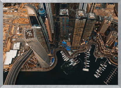 An aerial, high-angle photograph looking down on a modern city skyline, featuring towering skyscrapers, including a distinctive twisted tower, clustered around a dark-water marina filled with white yachts. A multi-lane highway snakes through the urban landscape, which has a warm, desaturated color palette and is enclosed in a silver frame. Decor