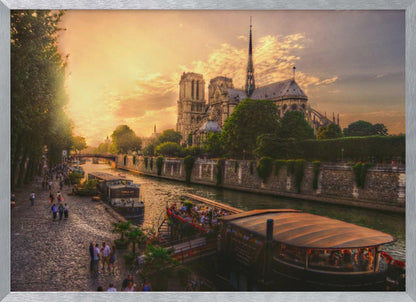 A scenic photograph of the Notre Dame Cathedral in Paris during a warm, golden sunset, as viewed from across the River Seine. People are strolling along the cobblestone riverbank and riding on tour boats docked on the water. The image is presented within a silver picture frame. Decor