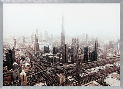 An aerial photograph of a sprawling modern cityscape on a hazy day, framed in silver. The iconic Burj Khalifa stands tall in the center background, surrounded by numerous other skyscrapers and a complex network of highways and interchanges in the foreground, all rendered in a muted, slightly sepia-toned color palette. Print