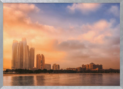 A framed photograph of the Abu Dhabi city skyline at sunset, featuring modern skyscrapers and the Emirates Palace hotel glowing in warm, orange light. The buildings are reflected in the calm water of the foreground, under a sky with soft orange and blue clouds. Poster